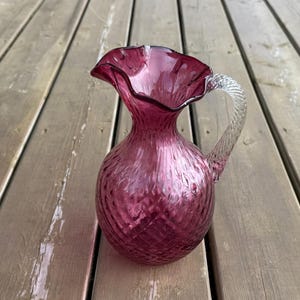 May include: A ruby-coloured glass jug with a ruffled rim and a clear, twisted handle. The jug has a textured, patterned surface. This elegant piece is suitable for serving beverages.