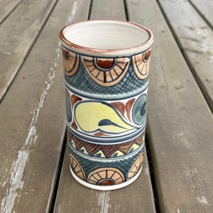 May include: A cylindrical ceramic vase with a white interior and a decorative exterior. The exterior features bands of repeating patterns in shades of brown, teal, yellow, and orange. The vase is approximately 15 cm tall.