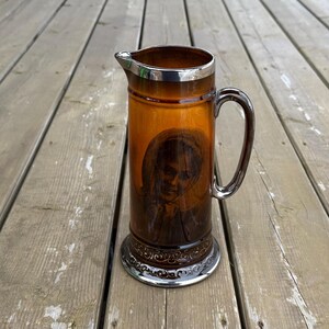 May include: Amber glass pitcher with silver-coloured rim, handle, and base. The pitcher features a portrait of a person. The handle is curved, and the base has a decorative pattern. A vintage decorative item.