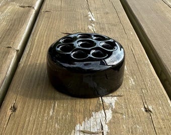 Op de afbeelding: Een ronde, glanzend zwarte keramische bloemenkikker met zeven cirkelvormige openingen in een patroon op het bovenoppervlak. Het object heeft een gladde, reflecterende afwerking en een cilindrische vorm met een platte basis.