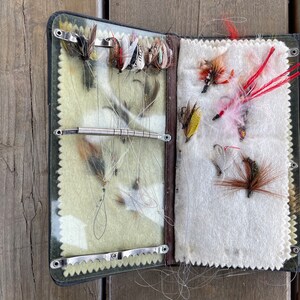 Puede incluir: Caja de aparejos de pesca con mosca abierta con una variedad de moscas artificiales. La caja es negra con un ribete verde y marr&oacute;n, y el interior tiene un forro de fieltro verde claro y blanco. Las moscas son de varios colores, incluyendo rojo, marr&oacute;n y blanco.