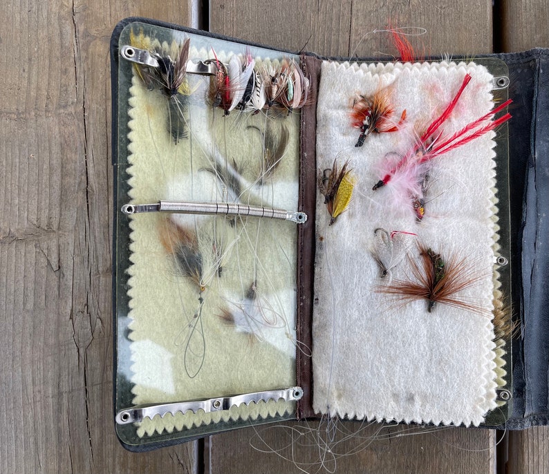 Puede incluir: Caja de aparejos de pesca con mosca abierta con una variedad de moscas artificiales. La caja es negra con un ribete verde y marr&oacute;n, y el interior tiene un forro de fieltro verde claro y blanco. Las moscas son de varios colores, incluyendo rojo, marr&oacute;n y blanco.