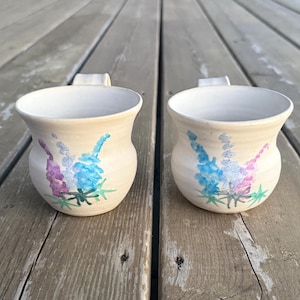 May include: Two cream-colored ceramic mugs with a floral design in blue, purple, and green. Each mug has a small handle and a wide opening. The mugs are sitting on a weathered wooden surface.