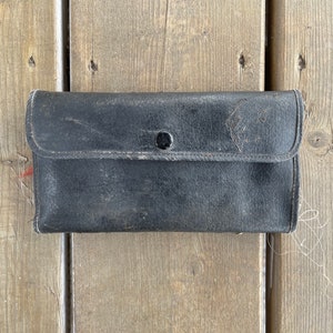 Puede incluir: Un monedero de cuero negro desgastado con cierre de bot&oacute;n. El monedero rectangular tiene una solapa y muestra signos de uso. Descansa sobre una superficie de madera envejecida.