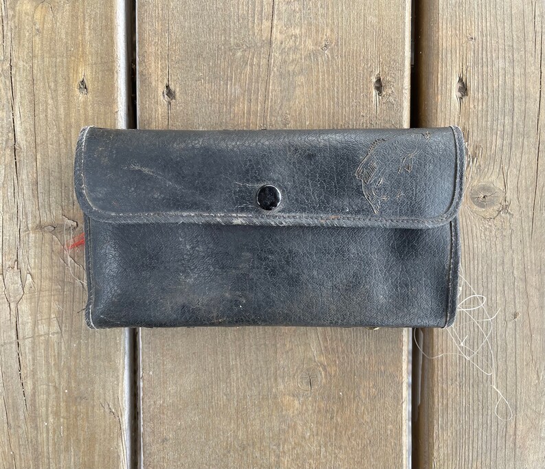Puede incluir: Un monedero de cuero negro desgastado con cierre de bot&oacute;n. El monedero rectangular tiene una solapa y muestra signos de uso. Descansa sobre una superficie de madera envejecida.