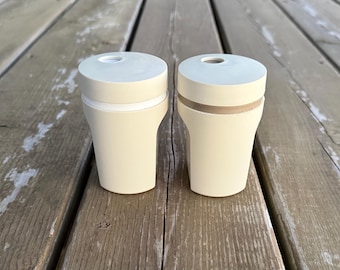 Op de afbeelding: Twee gebroken witte, cilindrische houders met platte, ronde deksels. Elk deksel heeft een kleine, ronde opening bij de rand. De houders hebben een gladde, matte afwerking en een licht taps toelopende vorm, naast elkaar geplaatst.