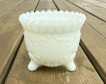 May include: A vintage, off-white, footed planter. The planter has a scalloped rim and is decorated with a floral pattern. The planter is made of a milky glass material and has three rounded feet.