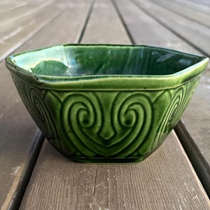 Peut inclure: Un bol en céramique vert avec un motif géométrique. Le bol a une forme carrée avec des coins arrondis et une finition brillante. L'extérieur présente un motif de tourbillon répétitif. Le bol est posé sur une surface en bois.