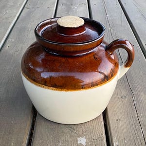 Peut inclure: Un pot en céramique émaillé brun et blanc avec un couvercle. Le pot a un émail brun avec une bande blanche autour du milieu. Le couvercle est également émaillé brun.