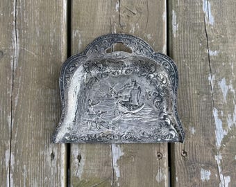 Op de afbeelding: Een zilverkleurige metalen kruimelschep met een reliëfscène van een zeilschip, een windmolen en figuren bij het water. De schep heeft een boogvormig handvat aan de bovenzijde en is versierd met gedetailleerde krulpatronen langs de rand.