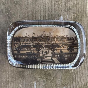May include: Rectangular glass ashtray featuring a sepia-toned image of a historical building and people. The ashtray has a clear, textured border. The image includes text that is difficult to read.
