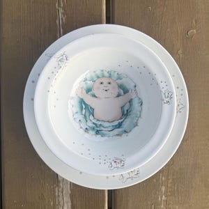 May include: A set of white ceramic plates and a bowl. The bowl has a baby illustration in a blue cabbage design. The plates have a dotted border with small bunny illustrations. The plates are on a wooden surface.