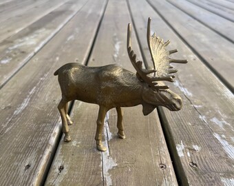 Può includere: Una statuetta di alce in ottone di profilo, caratterizzata da una finitura metallica dorata testurizzata, quattro zampe sottili e un grande set dettagliato di corna che si estendono dalla testa. L'animale è raffigurato in piedi con la testa bassa.