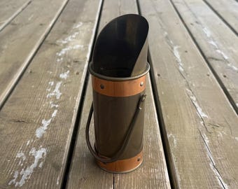 Può includere: Un secchio per carbone cilindrico in metallo con corpo color bronzo scuro e due fasce orizzontali color rame. Il contenitore presenta un'apertura superiore inclinata, un manico in metallo incernierato e rivetti visibili sulle fasce.