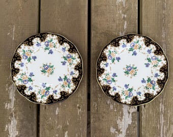 Può includere: Due piatti rotondi in porcellana con centro bianco e motivo floreale di fiori blu e gialli. I bordi smerlati sono decorati con un'ampia bordura blu navy scuro, accentuata da dettagli intricati in filigrana dorata.