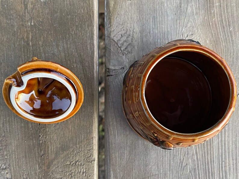 Op de afbeelding: Een bruine keramische pot met een deksel. De pot is versierd met een mandenweefpatroon en heeft een bruine glazuur. Het deksel is ook bruin en heeft een witte rand.