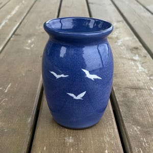 May include: A glossy, deep blue ceramic vase with a rounded body and a flared rim. The exterior surface features a speckled texture and is decorated with three small, white, stylized bird silhouettes arranged in a scattered pattern across the front.