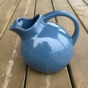 May include: A blue ceramic jug with a rounded body and a curved handle. The jug has a spout for pouring and is sitting on a wooden surface. The jug is approximately 20 cm tall.