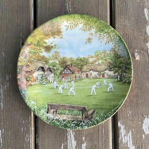 Peut inclure: Une assiette ronde en céramique avec une scène peinte d'un match de cricket de village. L'assiette représente un terrain vert avec des joueurs en uniforme blanc, un bâtiment au toit de chaume et un banc en bois. L'assiette a une bordure blanche avec un motif floral vert et blanc.