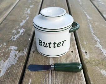 Op de afbeelding: Een witte keramische boterpot met een donkergroen handvat en deksel, voorzien van het woord Butter in donkergroene letters. Een bijpassend botermesje met een donkergroen handvat en een zilverkleurig metalen lemmet ligt voor de boterpot.