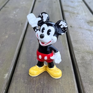 May include: A vintage Mickey Mouse figurine, with a black body, white gloves, and yellow shoes. The figure is waving with its right hand. The shorts are red with white buttons. The figurine is standing on a wooden surface.