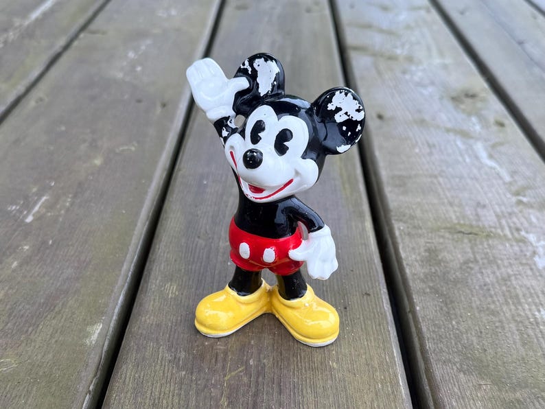 May include: A vintage Mickey Mouse figurine, with a black body, white gloves, and yellow shoes. The figure is waving with its right hand. The shorts are red with white buttons. The figurine is standing on a wooden surface.