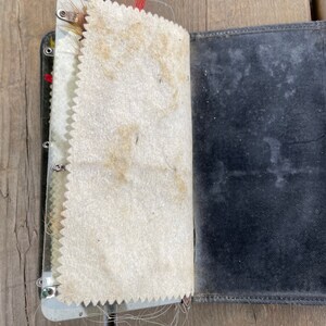 Puede incluir: Un diario de cuero negro vintage abierto, con lomo de metal y p&aacute;ginas color crema. Las p&aacute;ginas tienen un borde decorativo en zigzag. El diario muestra signos de desgaste y envejecimiento, con manchas visibles en las p&aacute;ginas y la cubierta.
