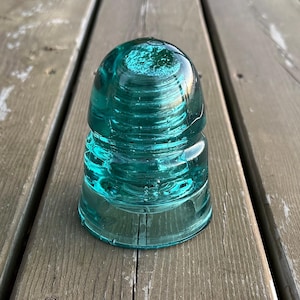 May include: A teal glass insulator with a rounded top and a ridged body. The glass has a slight swirl pattern and sits on a weathered wooden surface. A classic piece of industrial design, often used on telegraph poles.