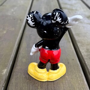 May include: A vintage Mickey Mouse figurine, with a black body, white accents on the ears, red shorts, white gloves, and yellow shoes. The figure is posed with one arm raised.