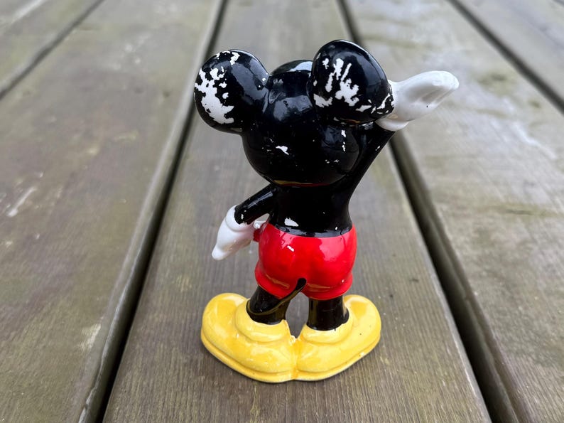 May include: A vintage Mickey Mouse figurine, with a black body, white accents on the ears, red shorts, white gloves, and yellow shoes. The figure is posed with one arm raised.