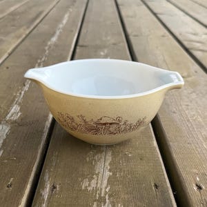 May include: A beige mixing bowl with a white interior and two pouring spouts. The exterior features a brown printed design of mushrooms and various wildflowers encircling the base. The bowl is constructed from glass and has a smooth, rounded shape.