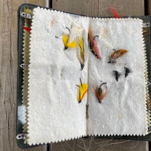 Puede incluir: Caja de aparejos de pesca con mosca abierta con varias moscas artificiales de colores. Las moscas est&aacute;n adheridas a un interior de fieltro blanco. La caja es negra con un cierre met&aacute;lico y est&aacute; abierta sobre una superficie de madera.