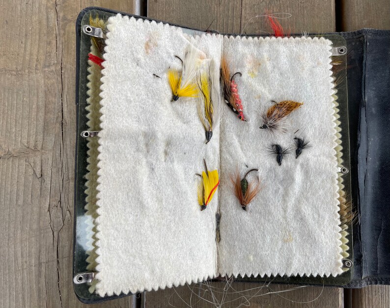 Puede incluir: Caja de aparejos de pesca con mosca abierta con varias moscas artificiales de colores. Las moscas est&aacute;n adheridas a un interior de fieltro blanco. La caja es negra con un cierre met&aacute;lico y est&aacute; abierta sobre una superficie de madera.