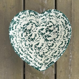 Peut inclure: Bol en forme de cœur avec un motif tacheté blanc et vert foncé. Le bol est en céramique ou en émail et repose sur une surface en bois. Le design du bol rappelle la vaisselle émaillée vintage.