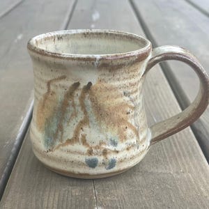 May include: A handmade ceramic mug with a brown, white and blue glaze. The mug has a rounded body and a handle.