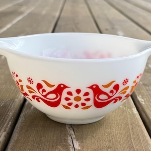 May include: White mixing bowl with a decorative red and orange bird and floral pattern. The bowl has two handles and is set on a wooden surface. A vintage kitchenware item.
