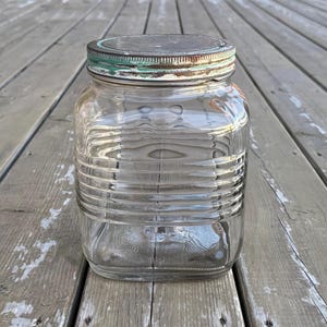 May include: A clear, square glass jar with horizontal ribbed detailing. The jar has a metal lid with a green and rust-coloured finish. The jar is on a weathered wooden surface.