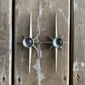 May include: Two identical silver-toned metal starburst wall ornaments. Each piece features a central circular concave element surrounded by eight radiating pointed spikes of varying lengths, creating a symmetrical geometric design.