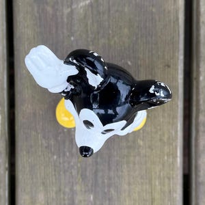 May include: A black, white, and yellow Mickey Mouse figurine. The figurine has a glossy finish with some paint wear. The ears are black, the face is white, and the gloves are white. The figurine is on a wooden surface.