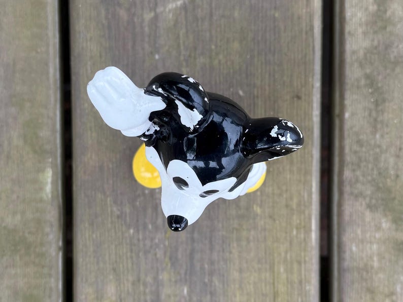 May include: A black, white, and yellow Mickey Mouse figurine. The figurine has a glossy finish with some paint wear. The ears are black, the face is white, and the gloves are white. The figurine is on a wooden surface.