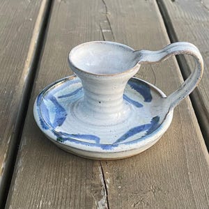 Peut inclure: Un bougeoir en céramique blanche avec un motif bleu et blanc. Le bougeoir a une poignée et un petit plat pour la bougie.