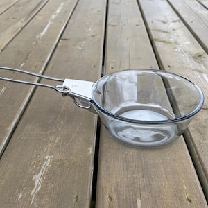 May include: A clear glass bowl with a metal handle attached. The bowl has a dark blue rim and is set against a wooden background. The handle is made of a thin, silver-coloured metal.