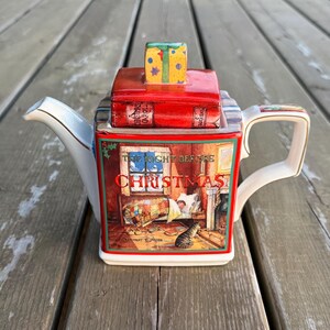 May include: A ceramic teapot shaped like a stack of books with a red lid featuring a small gift box topper. The front panel displays an illustration titled The Night Before Christmas, showing a bedroom scene with a cat, a bed, and a window.