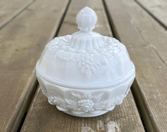Può includere: Una zuccheriera in vetro opalino bianco con coperchio abbinato, caratterizzata da un motivo in rilievo di uva e foglie. Il contenitore circolare presenta una base sfaccettata e un coperchio sormontato da un pomello appuntito.