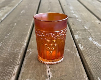 Op de afbeelding: Een oranje iriserende glazen beker met een geperst patroon van druiventrossen en bladeren rond de bovenkant. Het glas heeft een gladde rand en een cilindrisch lichaam dat iets taps toeloopt naar de basis toe.