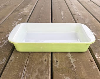 Può includere: Una pirofila rettangolare con un esterno liscio color verde chiaro e un interno bianco pulito. Il recipiente presenta manici integrati su entrambe le estremità corte, una base piatta e pareti leggermente svasate in materiale lucido.