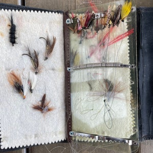 Puede incluir: Una caja de aparejos de pesca con mosca vintage abierta. La caja es de cuero negro y tiene una cubierta de pl&aacute;stico transparente. En el interior, varias moscas de pesca coloridas est&aacute;n dispuestas sobre un respaldo de fieltro blanco. Las moscas son de varios colores, incluyendo rojo, amarillo y marr&oacute;n.