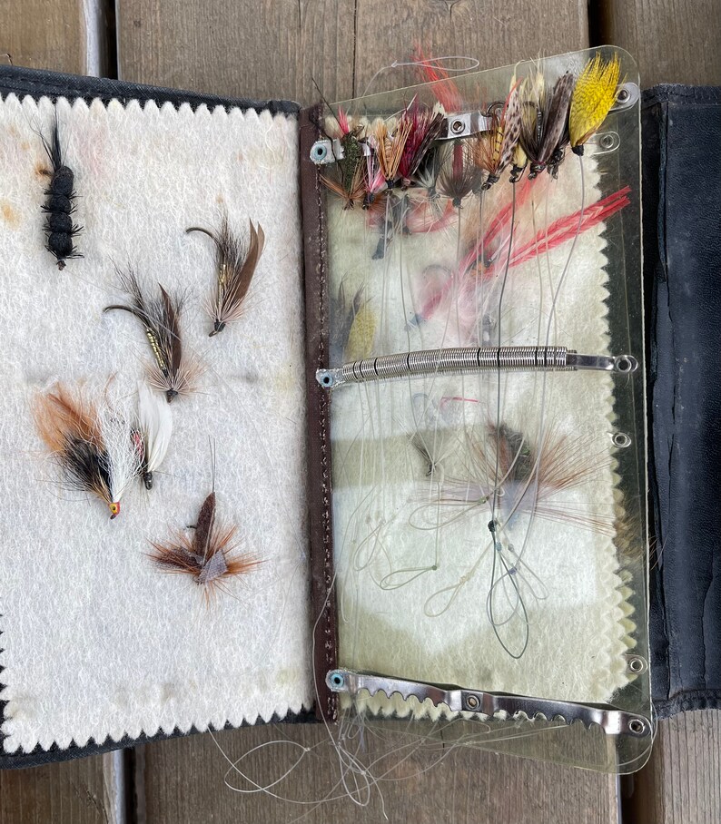 Puede incluir: Una caja de aparejos de pesca con mosca vintage abierta. La caja es de cuero negro y tiene una cubierta de pl&aacute;stico transparente. En el interior, varias moscas de pesca coloridas est&aacute;n dispuestas sobre un respaldo de fieltro blanco. Las moscas son de varios colores, incluyendo rojo, amarillo y marr&oacute;n.