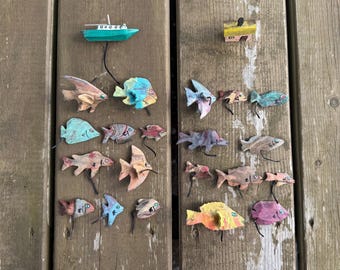Può includere: Una collezione di piccoli ornamenti in legno a forma di pesce, dipinti a mano in varie forme e colori, tra cui tonalità di blu, rosa, marrone e giallo. Sono inclusi una piccola barca verde e una casa gialla, ciascuna con un cordino nero.
