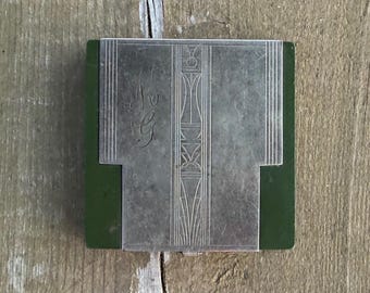 Può includere: Un astuccio compatto quadrato con una base verde scuro e un rivestimento in metallo color argento. La superficie metallica presenta linee geometriche incise e un pannello centrale verticale con motivi stilizzati, oltre a un monogramma inciso MG.